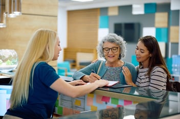Elderly woman and her daughter speaking with a GP receptionist while filling out a form, representing how EMMA by QuantumLoopAI helps overcome language barriers and patient frustration in NHS GP bookings.