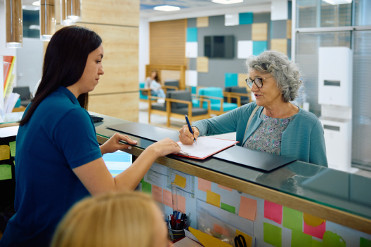 Elderly woman signing paperwork at a GP surgery reception desk, representing how EMMA by QuantumLoopAI supports NHS practices by reducing admin pressure and improving patient access through AI call handling.