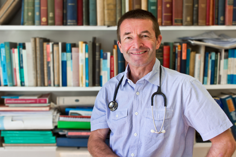 Smiling male GP wearing a stethoscope standing in front of a bookshelf, representing how EMMA by QuantumLoopAI supports clinicians in NHS GP surgeries by improving efficiency and patient communication through AI reception.