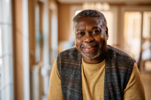 Smiling man sitting indoors wearing a brown sweater and patterned vest, representing how EMMA by QuantumLoopAI helps NHS GP surgery patients get through on the first call to manage prescriptions and appointments without stress.