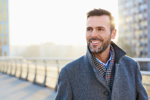 Smiling man wearing a grey coat standing outdoors on a bridge, representing how EMMA by QuantumLoopAI provides NHS GP surgery patients with privacy to discuss sensitive health concerns over the phone without embarrassment or judgment.