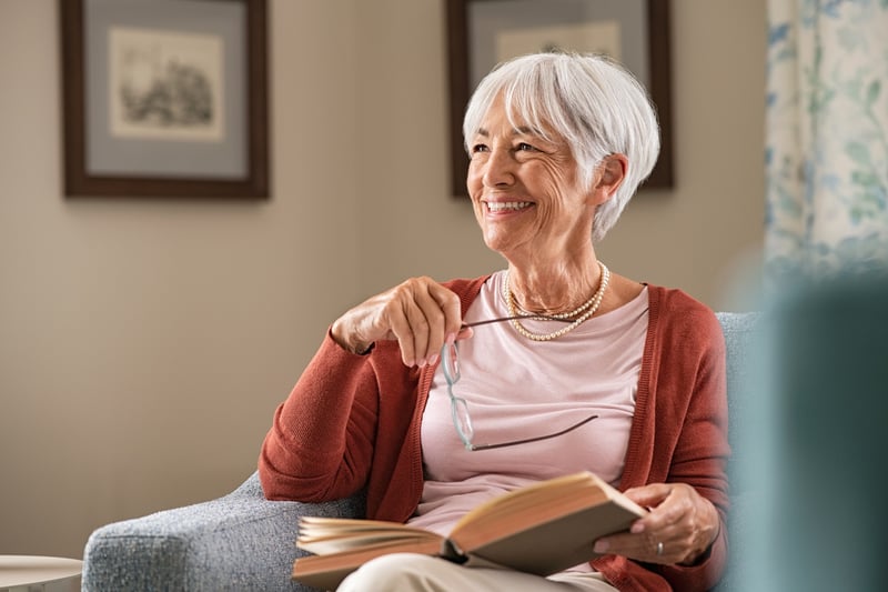 Smiling older woman sitting on a sofa holding a book and glasses, representing a relaxed patient enjoying easier access to healthcare through EMMA by QuantumLoopAI, the AI reception for NHS GP surgeries.