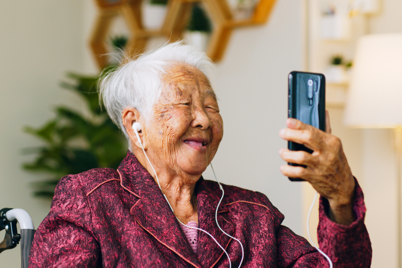 Smiling elderly woman using a smartphone with earphones during a video call, representing how EMMA by QuantumLoopAI helps NHS GP surgery patients communicate easily and access care through AI reception technology.